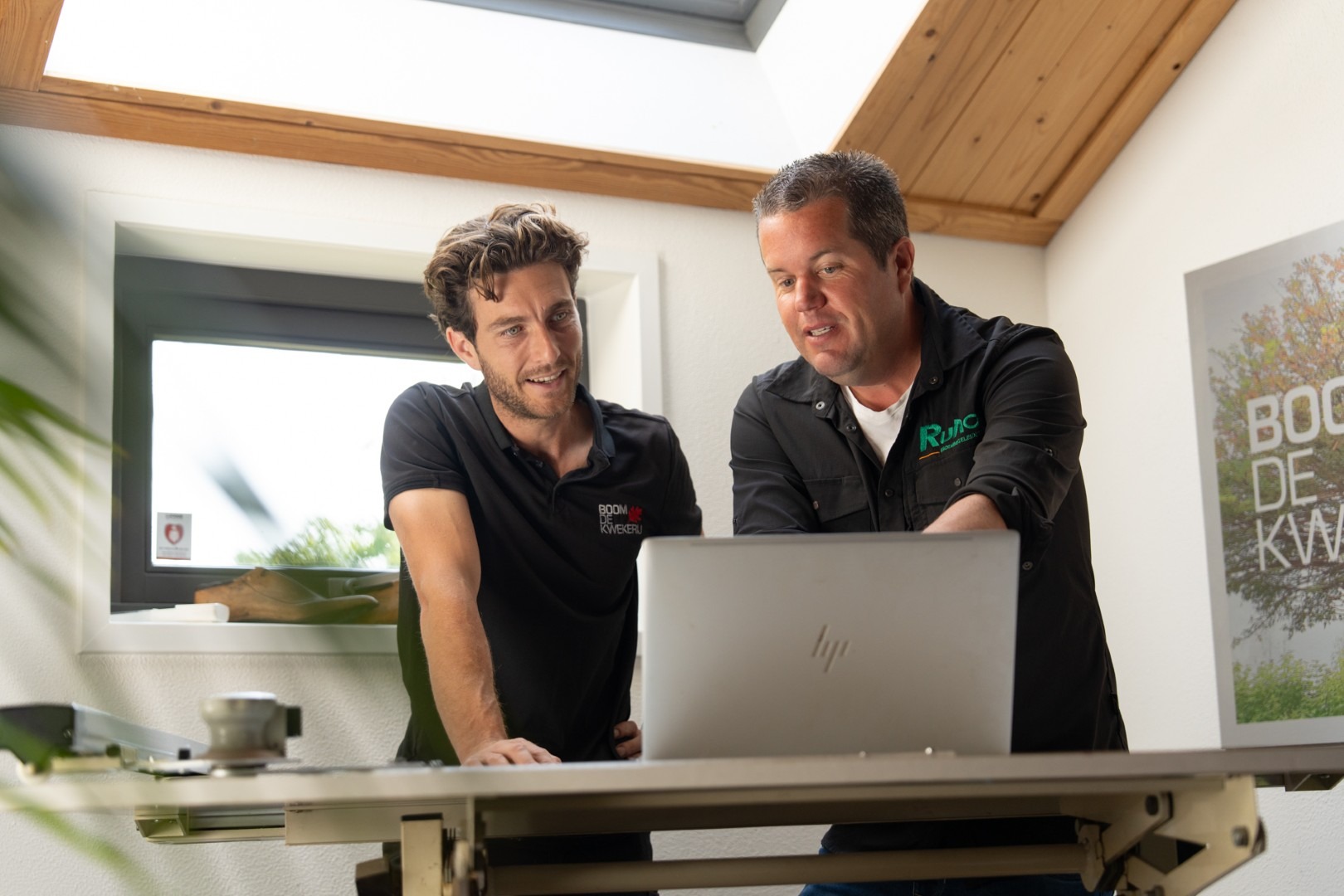 Rutger Hornikx van Ruho Groenbegeleiding staat binnen achter een in hoogte verstelbaar bureau, draagt een zwart overhemd met het Ruho-logo en wijst iets aan op het scherm van een laptop, terwijl een andere man geïnteresseerd meekijkt.